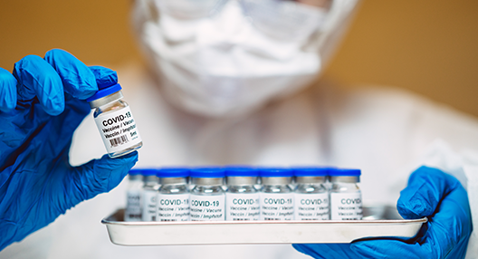 Slightly out-of-focus close-up of a white tray with several small glass vials with labels and a white cap that appear to be vaccines. The tray is held in the left hand of someone wearing blue gloves. In the foreground, and completely in focus, the person’s right hand holds a vial labelled “Covid-19 vaccine” and which looks similar to those on the tray.